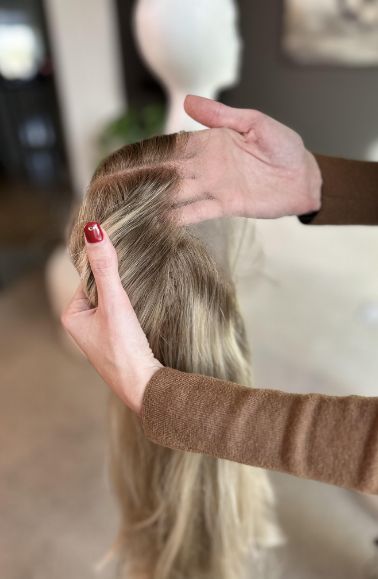 A person with red nail polish holds a long, blonde wig by the hairline, showing the lace front of a Medical Grade Wig. The background is softly blurred in neutral tones, with an indistinct mannequin head visible.